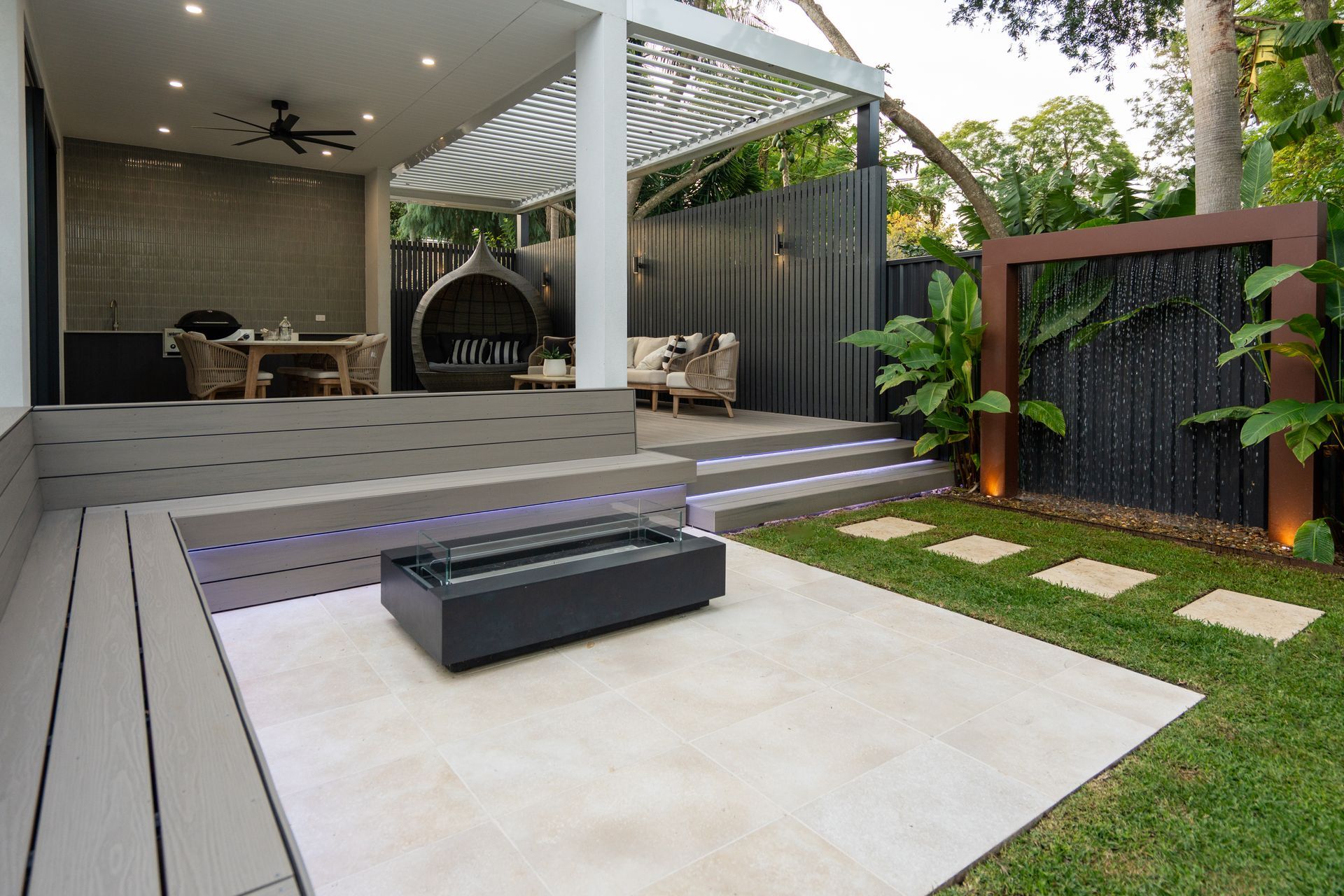 There is a fire pit in the middle of the backyard, surrounded by the outdoor kitchen and dining area and water feature.