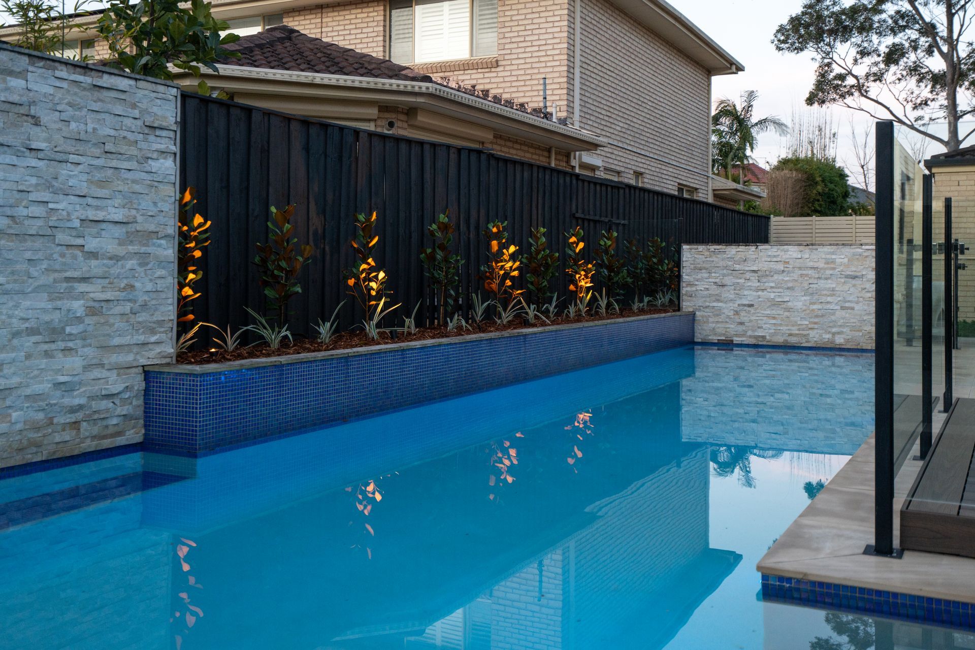 Pool with blue water and tiled edges, framed by a stone wall, black fence, and illuminated plants.