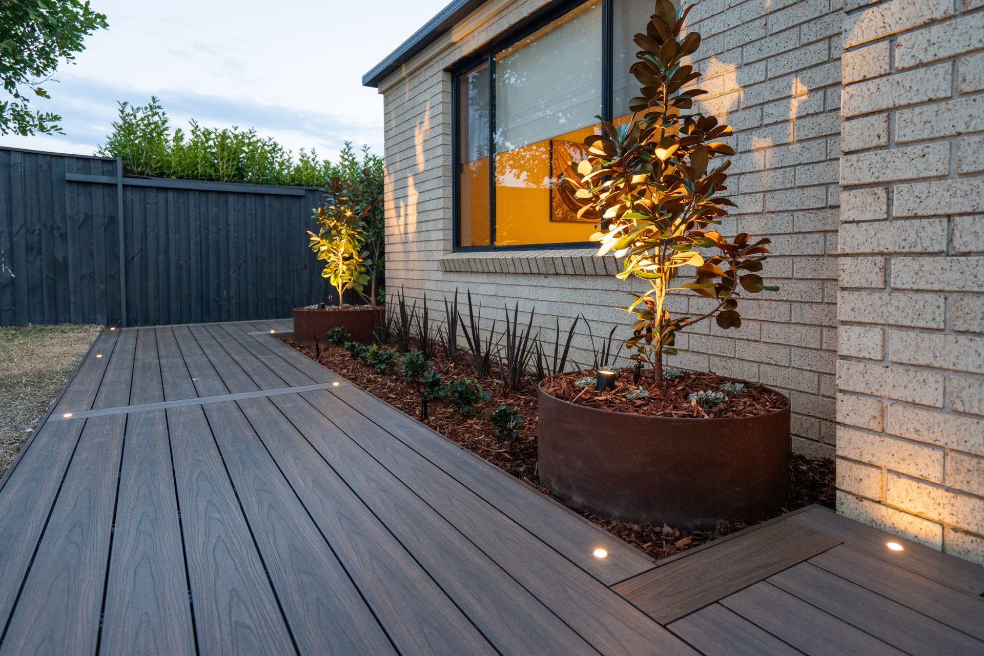 Composite deck pathway with recessed lights next to a brick wall with planters and trees, lit up at dusk.