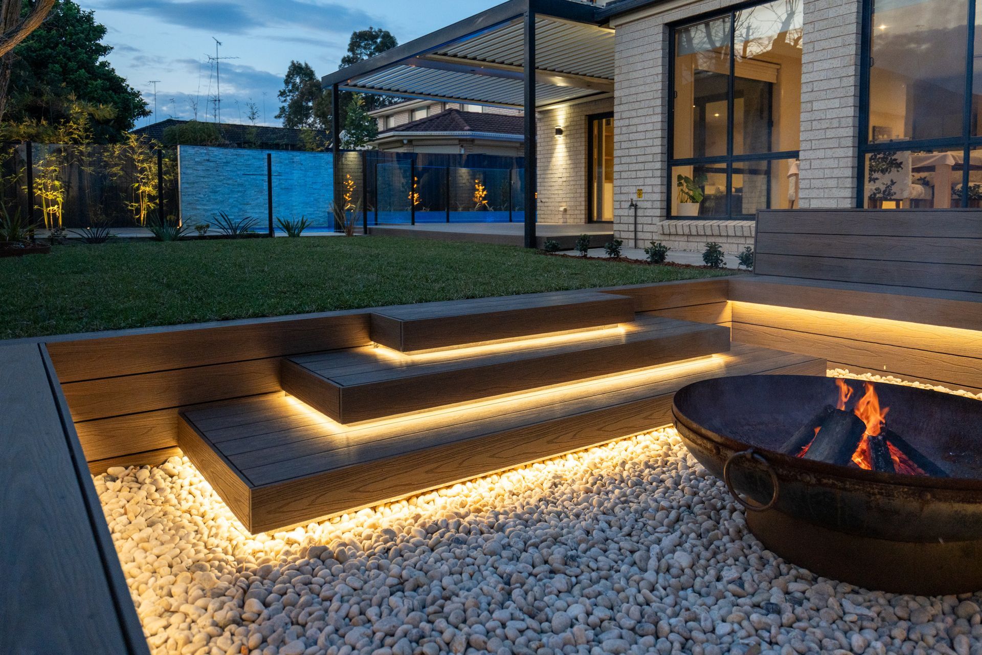 Backyard with steps lit by warm lights, fire pit on gravel, and modern house in the background.
