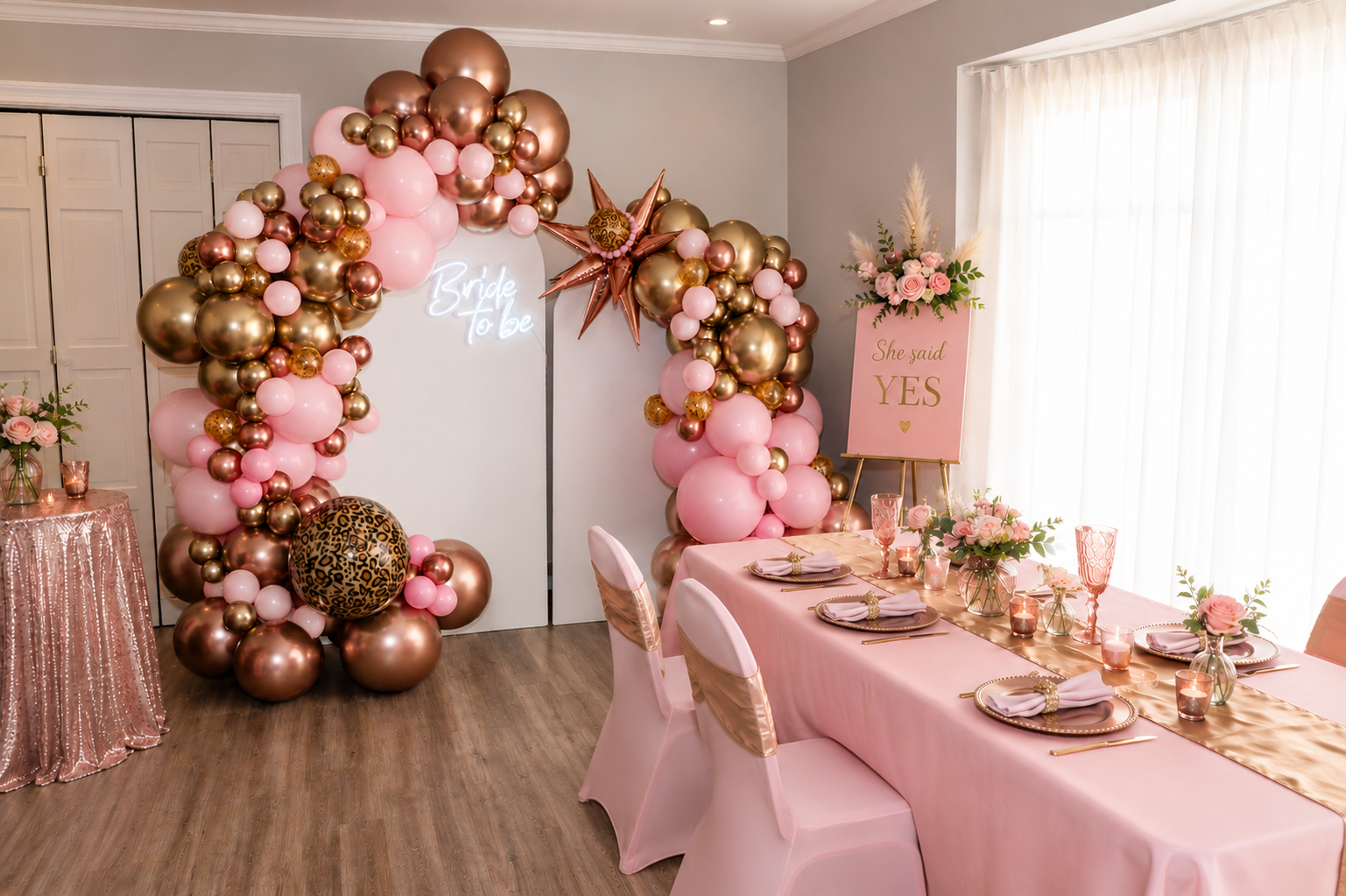 Pink-and-gold balloon arch beside a decorated birthday table in a bright party room