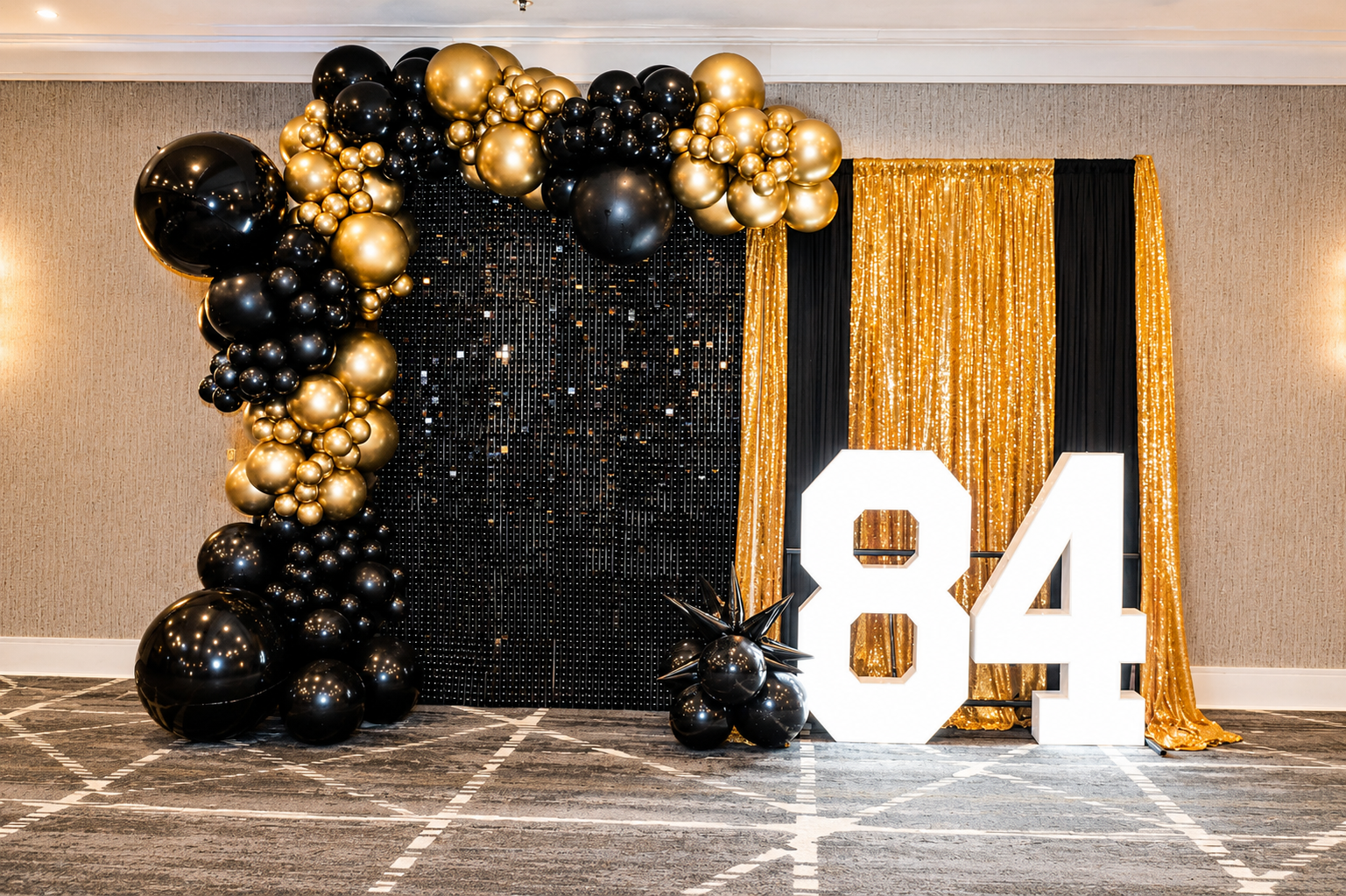 Black and gold balloon display with white “84” numbers and gold fringe backdrop at an event