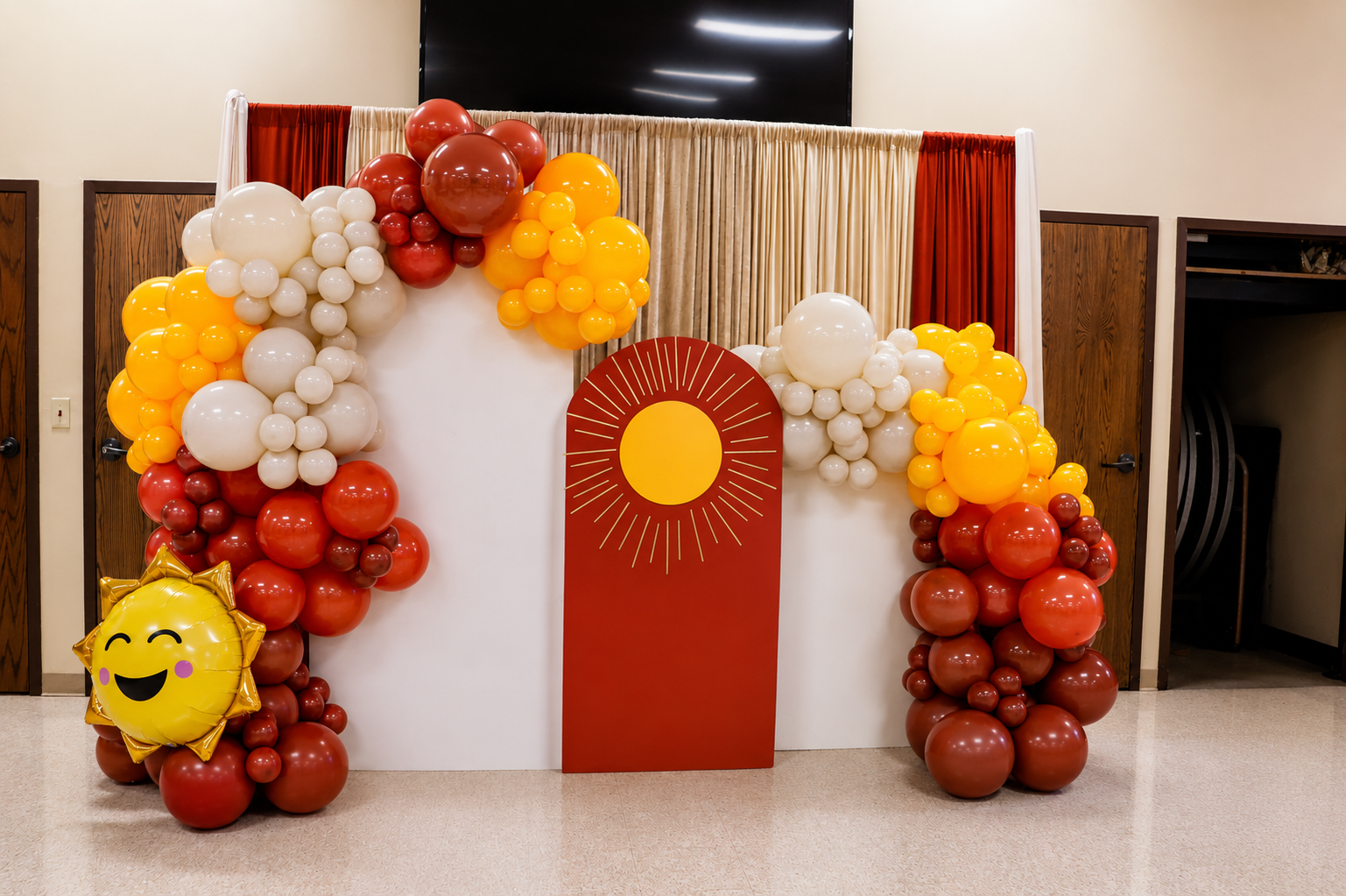 Red, white, and yellow balloon arch with sun decoration and emoji balloon in an indoor event setup