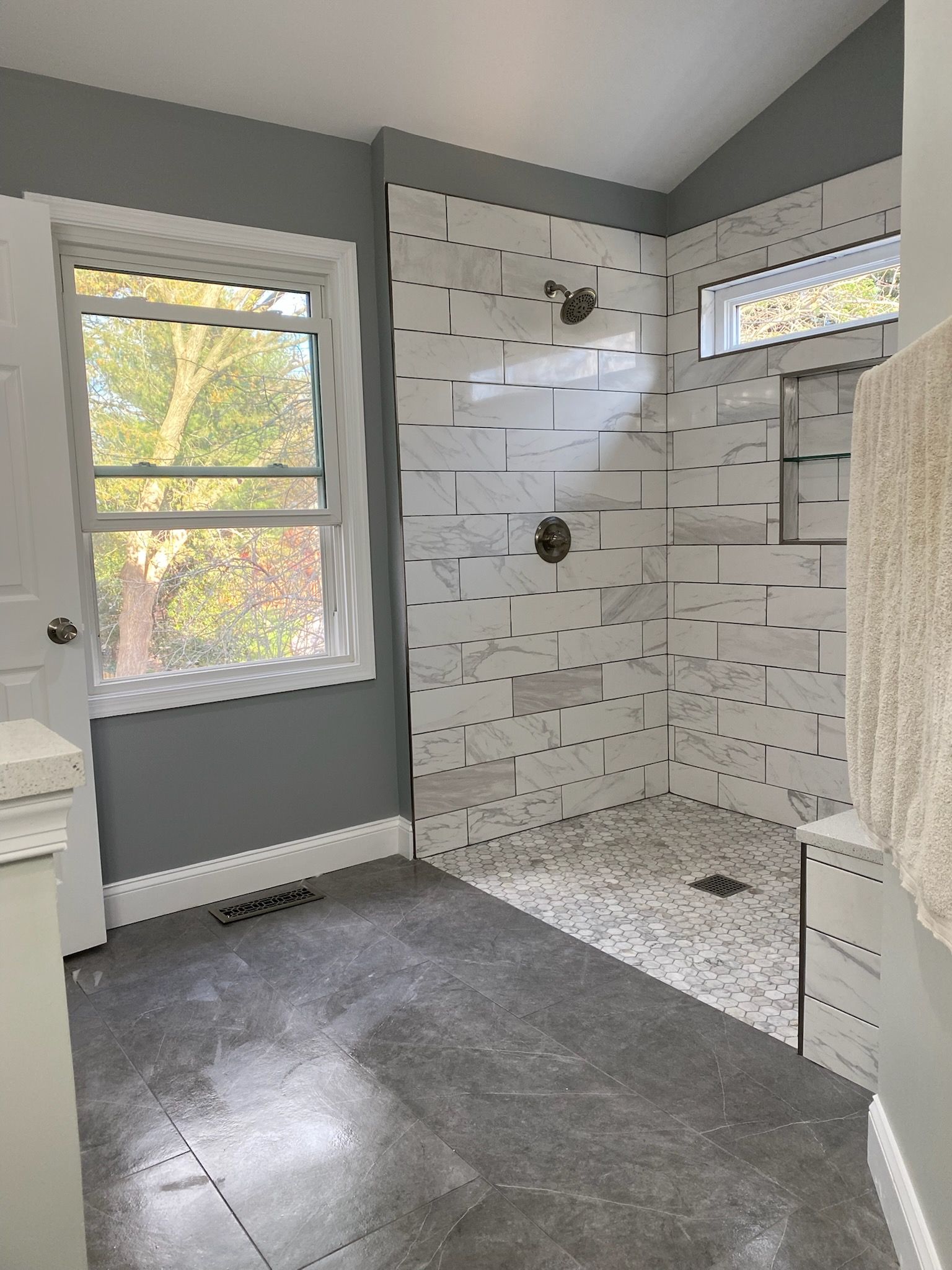 Bright bathroom with gray walls, white tile shower, window, and gray floor, lit by natural light