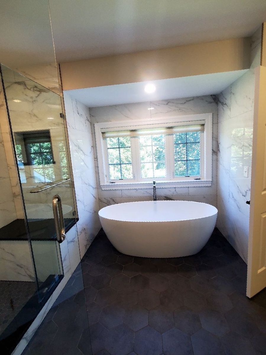 Modern bathroom with freestanding oval tub, large window, dark floor, and glass shower on the left