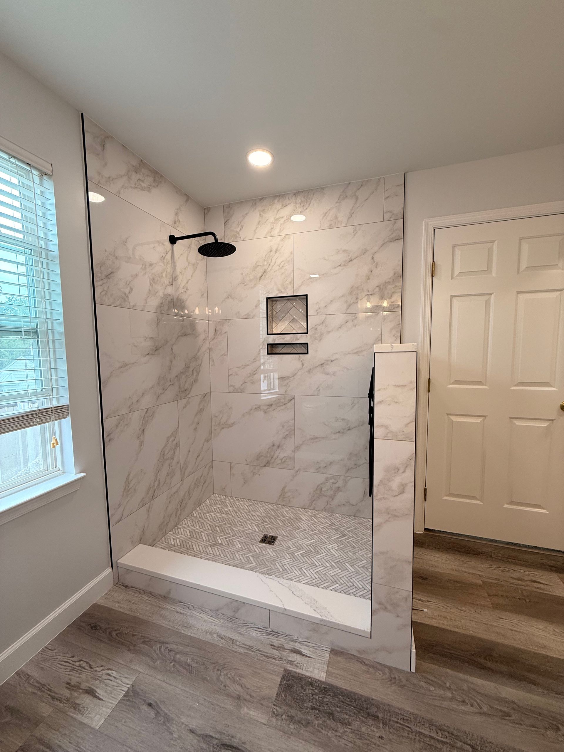 Marble-tiled walk-in shower with black showerhead, bench, and white door in a bright bathroom
