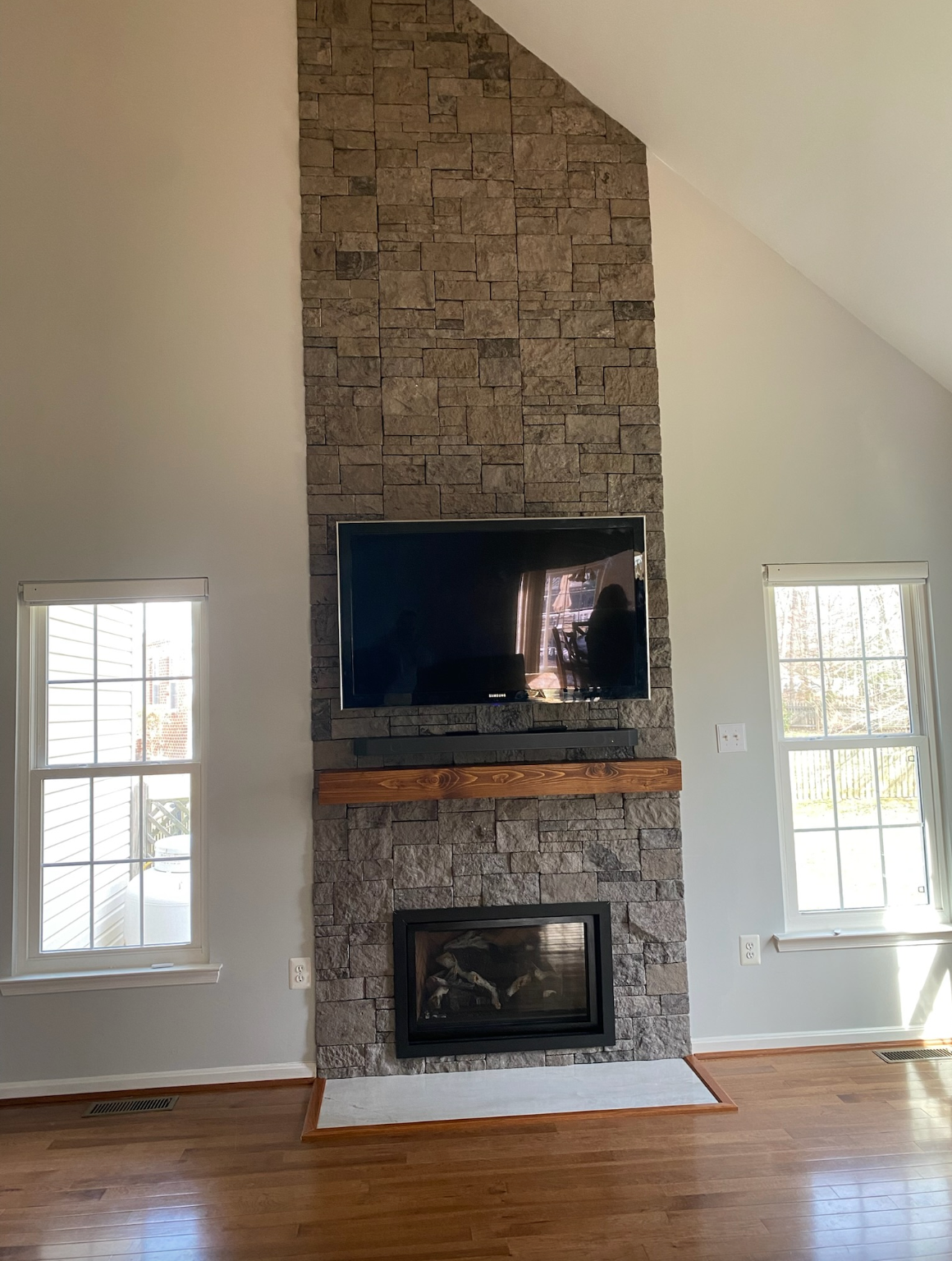 Living room with a stone fireplace, two windows, and hardwood floors