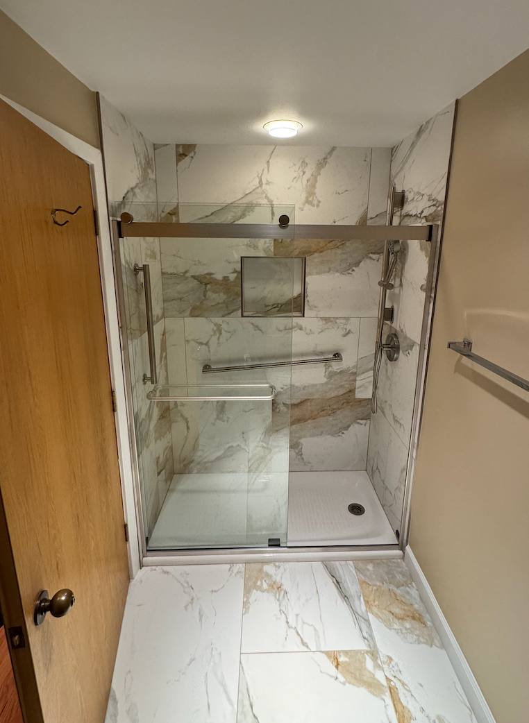 Small marble-tiled shower stall in a narrow bathroom with glass doors and a chrome towel bar.
