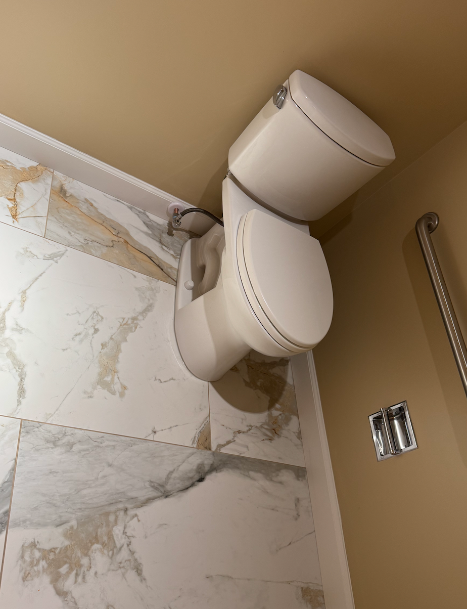 Tilted white wall-mounted toilet beside marble tile and tan wall in a bathroom