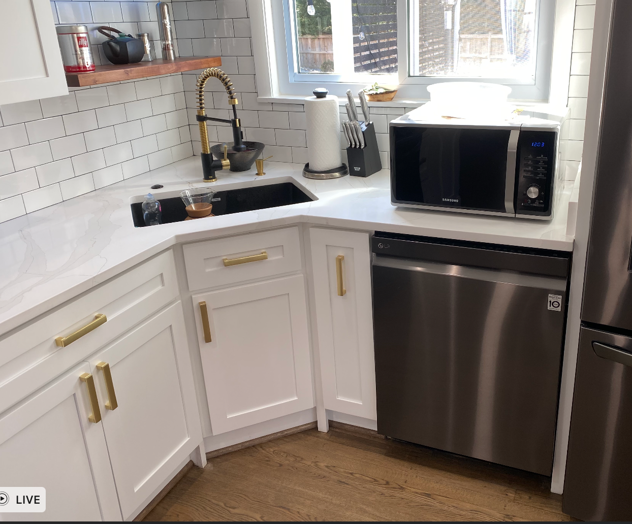 Modern white kitchen corner with black countertop, stainless dishwasher, sink, and microwave by a window.