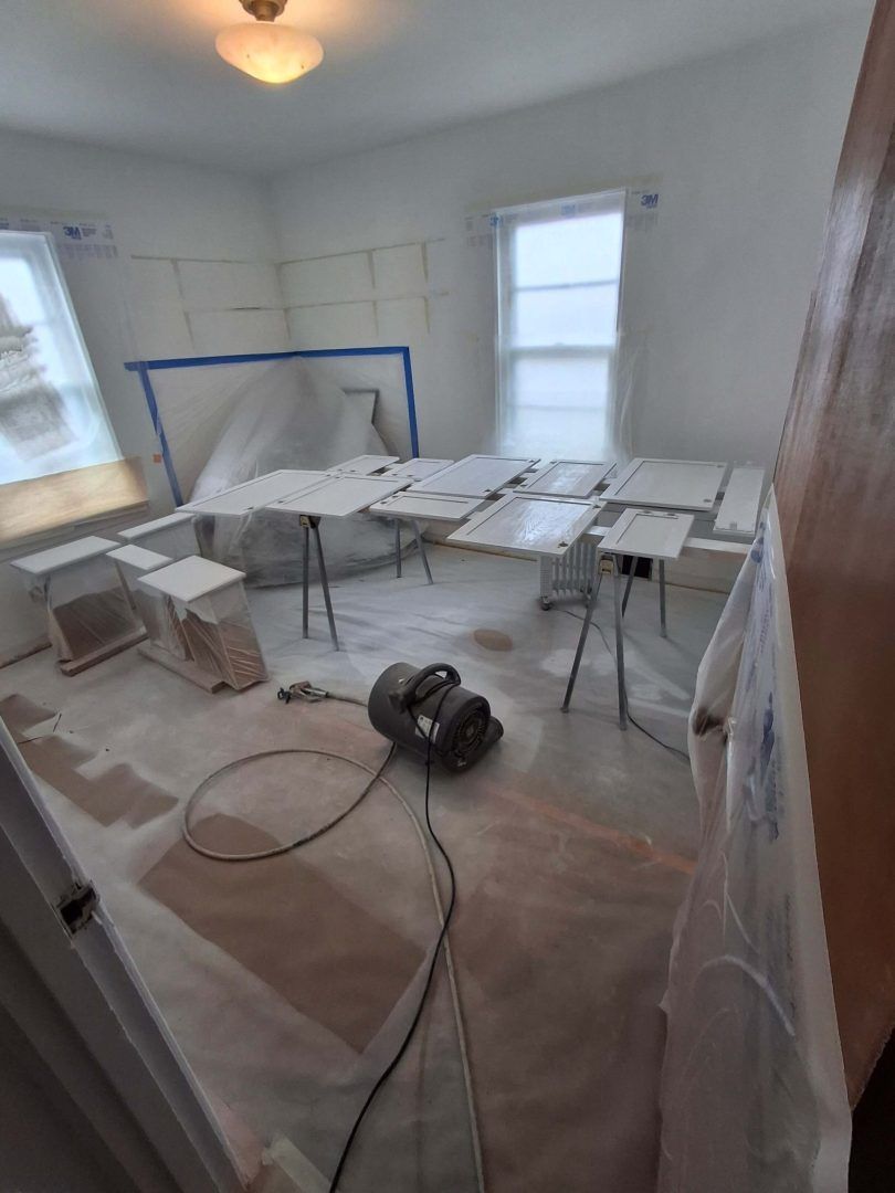 Painted room under renovation with drop cloths, taped trim, folding tables, and a floor fan. Painted room under renovation with drop cloths, taped trim, folding tables, and a floor fan.