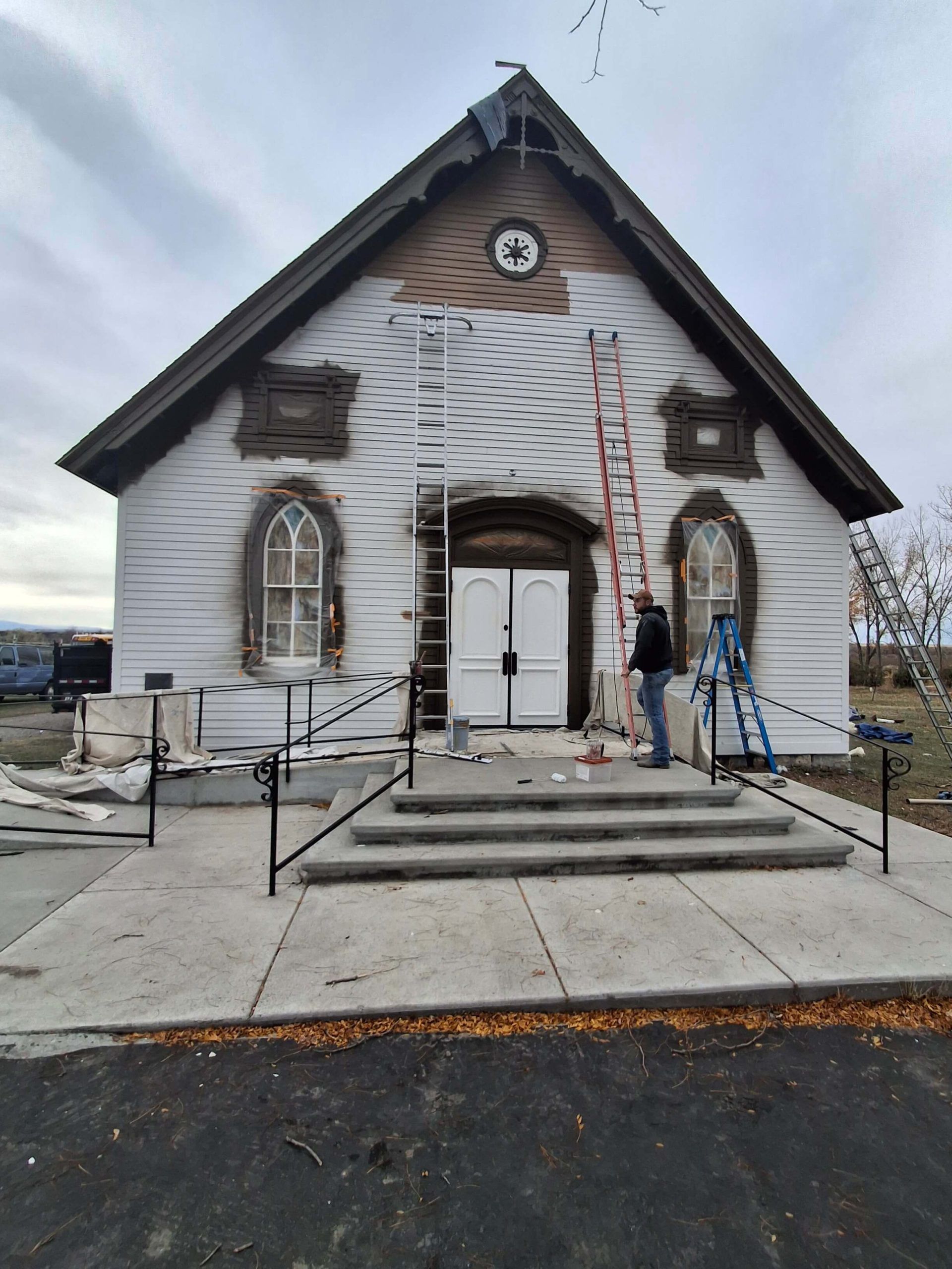 White church facade with arched windows, cross ladder, and two workers on steps under overcast sky White church facade with arched windows, cross ladder, and two workers on steps under overcast sky
