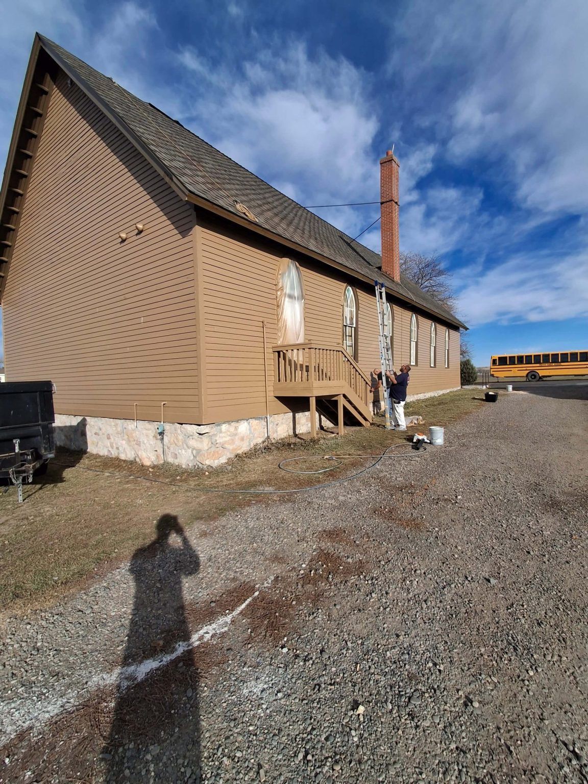 Brown wooden building on gravel under a blue sky, with a person near the entrance and a shadow in the foreground