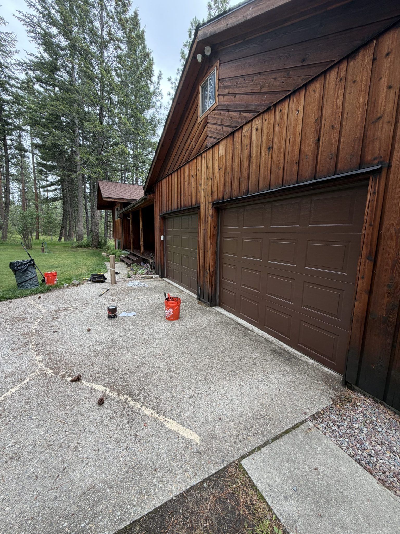 Gravel driveway beside a brown wooden house and garage, with trees and a small black trash bin nearby. Gravel driveway beside a brown wooden house and garage, with trees and a small black trash bin nearby.