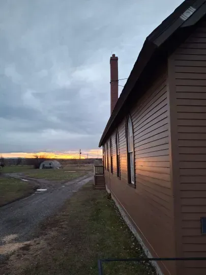 Side of a house at dusk beside a dirt road, with a cloudy sky and glowing sunset on the horizon.