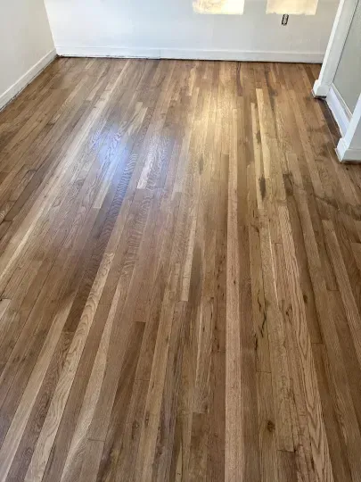 Empty room with glossy wood flooring and white walls, lit by sunlight from a window.