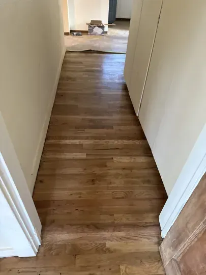 Narrow hallway with wood floor and white walls leading to a brighter room at the end