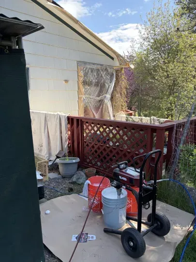 Backyard beside a white house with a red lattice fence and a wheelbarrow, buckets, and tools on the ground