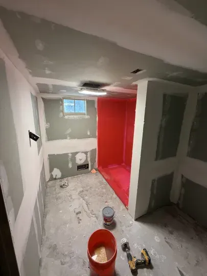 Small unfinished bathroom with red shower area, gray drywall, window, and paint buckets on the floor