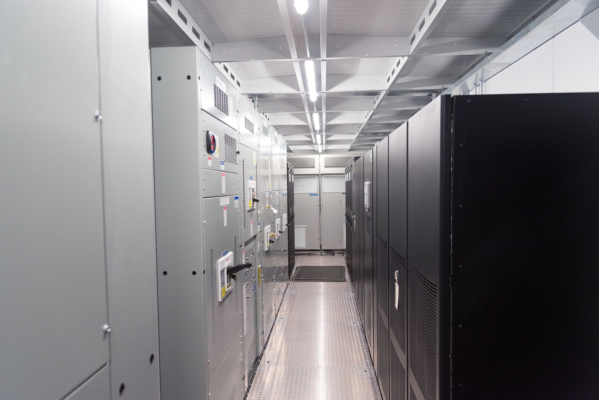 A narrow server room aisle with rows of black and gray racks and bright overhead lighting.