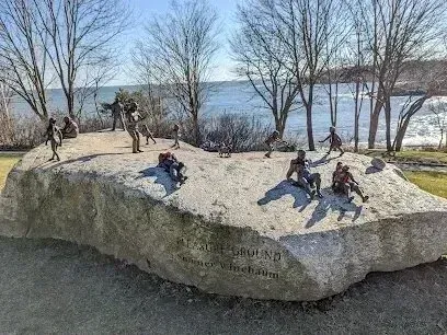 Chimney-Sweep-Portland-Bronze sculptures of people are scattered across a large, flat rock with carved text, set against a backdrop of trees and a body of water.