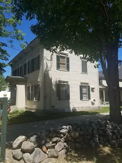 Chimney-Sweep-Portland-A light-colored, two-story clapboard house with dark shutters stands behind a low stone retaining wall under the shade of a large tree against a bright blue sky.