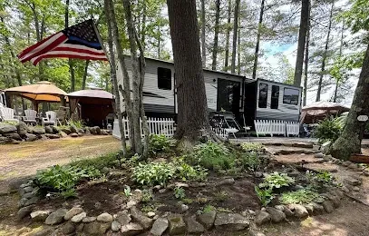 Chimney-Sweep-Portland-Camping scene with a travel trailer surrounded by tall trees and a garden with a small rock border. An American flag flies, creating a peaceful, rustic atmosphere.