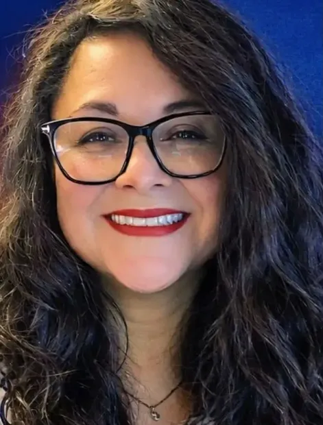 Smiling person with black glasses and wavy dark hair against a blue background