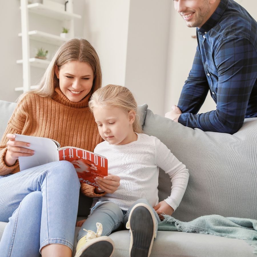 Family reading together on a couch, with a woman and child looking at a book while a man leans in behind them