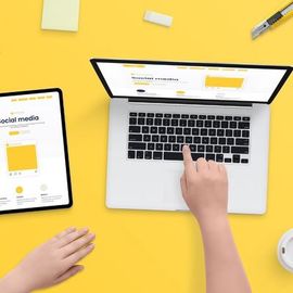 Top view of hands using a laptop and tablet on a bright yellow desk with a coffee cup and stationery