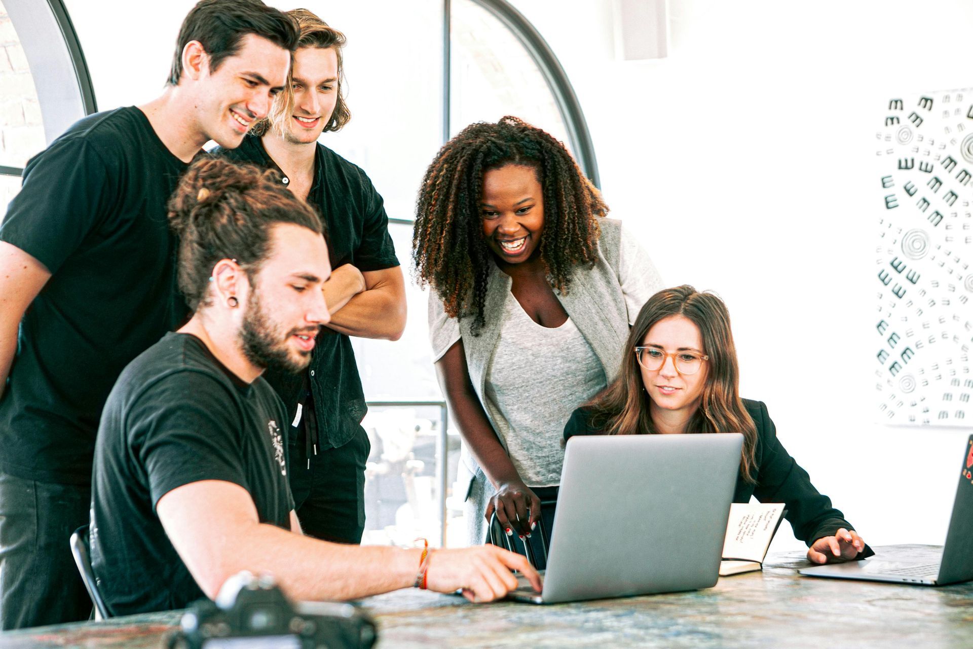 Five diverse people gathered around a laptop, collaborating in a bright office, smiling.