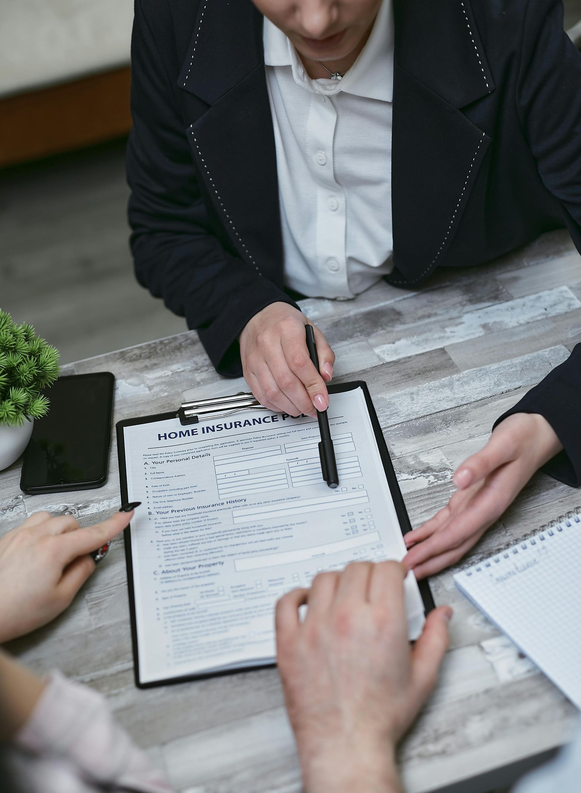 Person in a suit pointing at a contract on a clipboard; two people seated at a table.