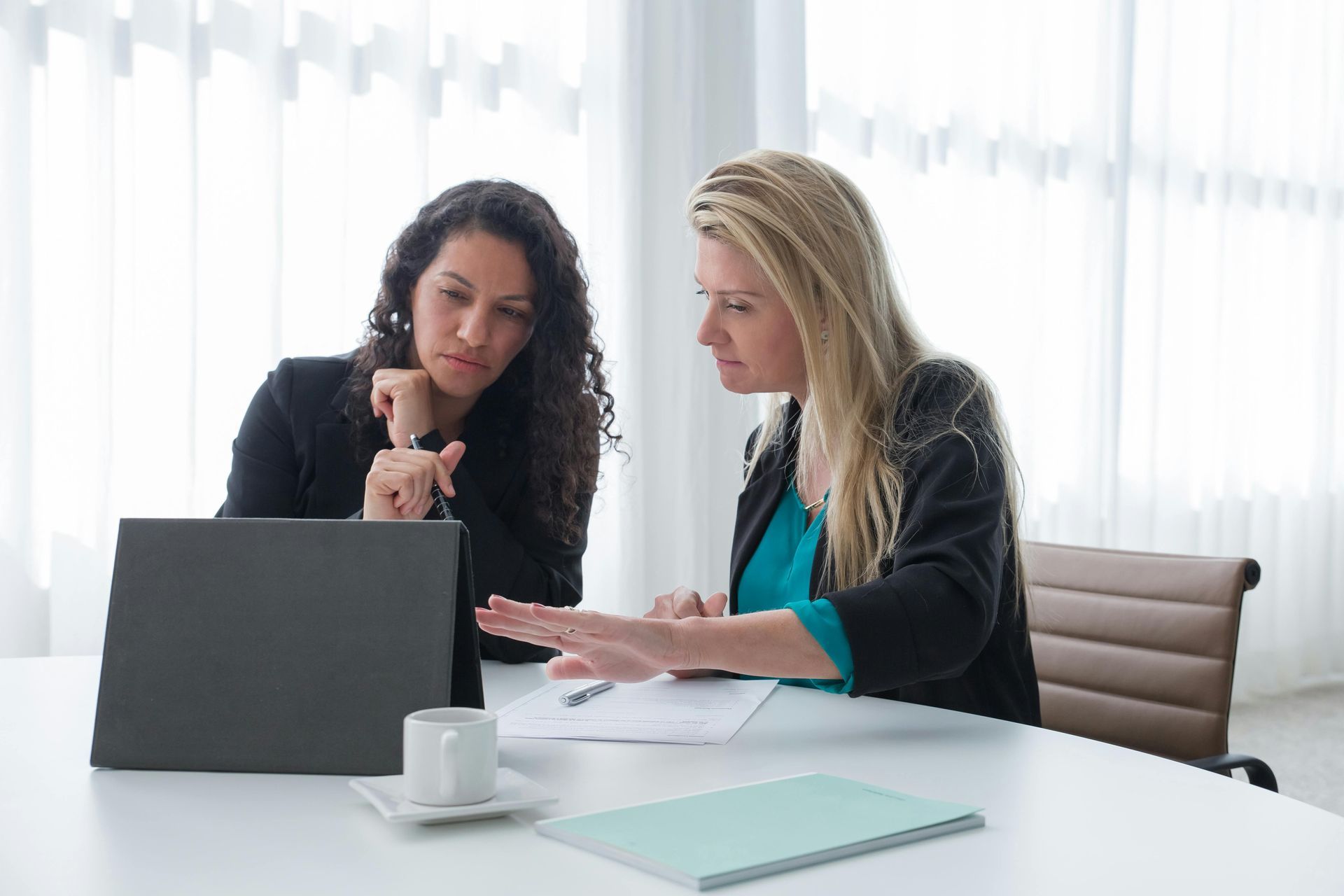 Two women at a table reviewing papers and a laptop. One points at a document.