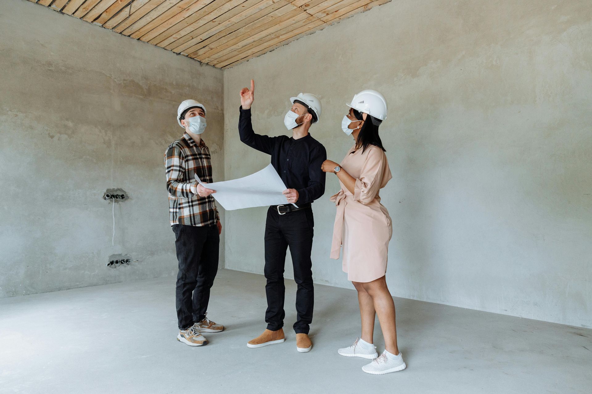 Three people in hard hats and masks review plans in a construction site.