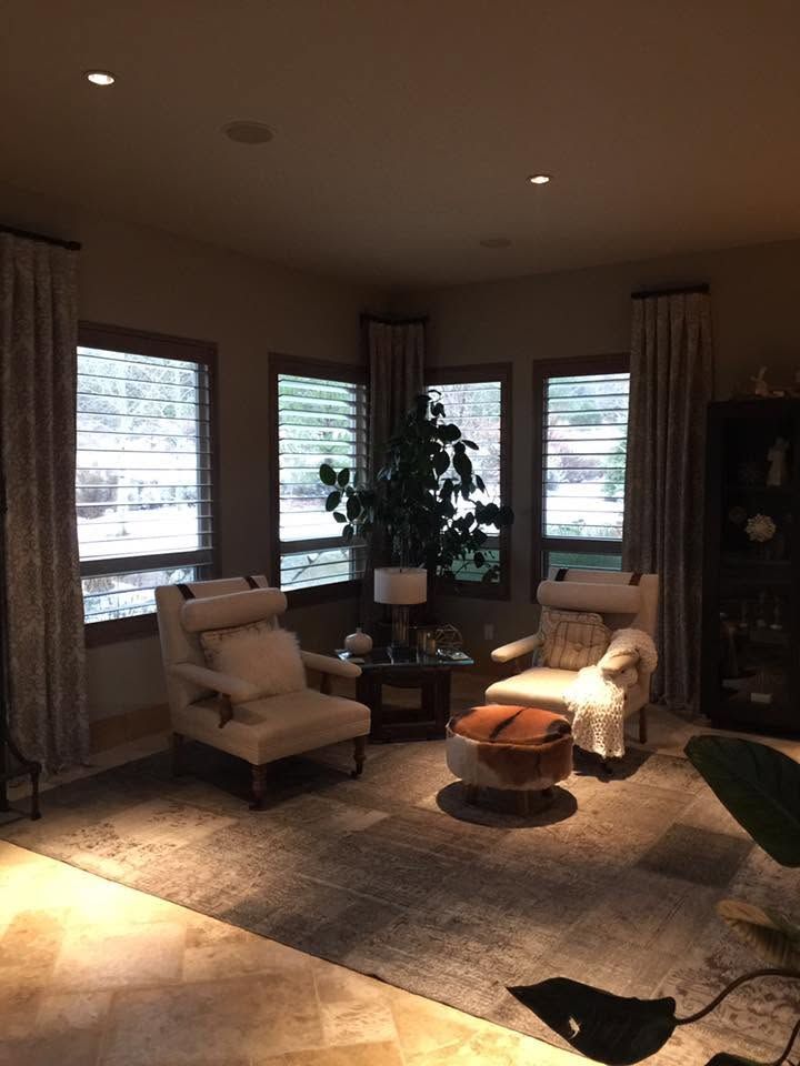 Cozy living room with armchairs, round ottoman, large windows, and warm sunlight on a rug.