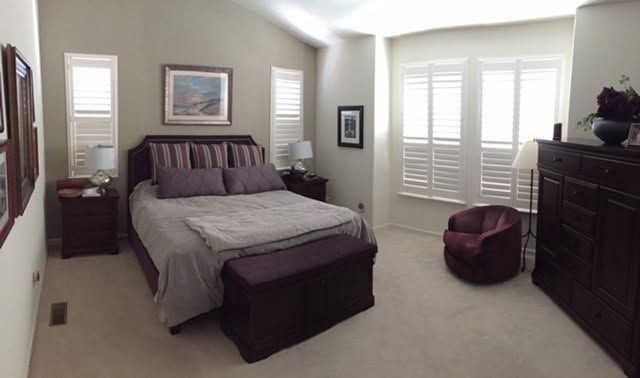Bright bedroom with a bed, dresser, windows with shutters, and a brown ottoman chair.