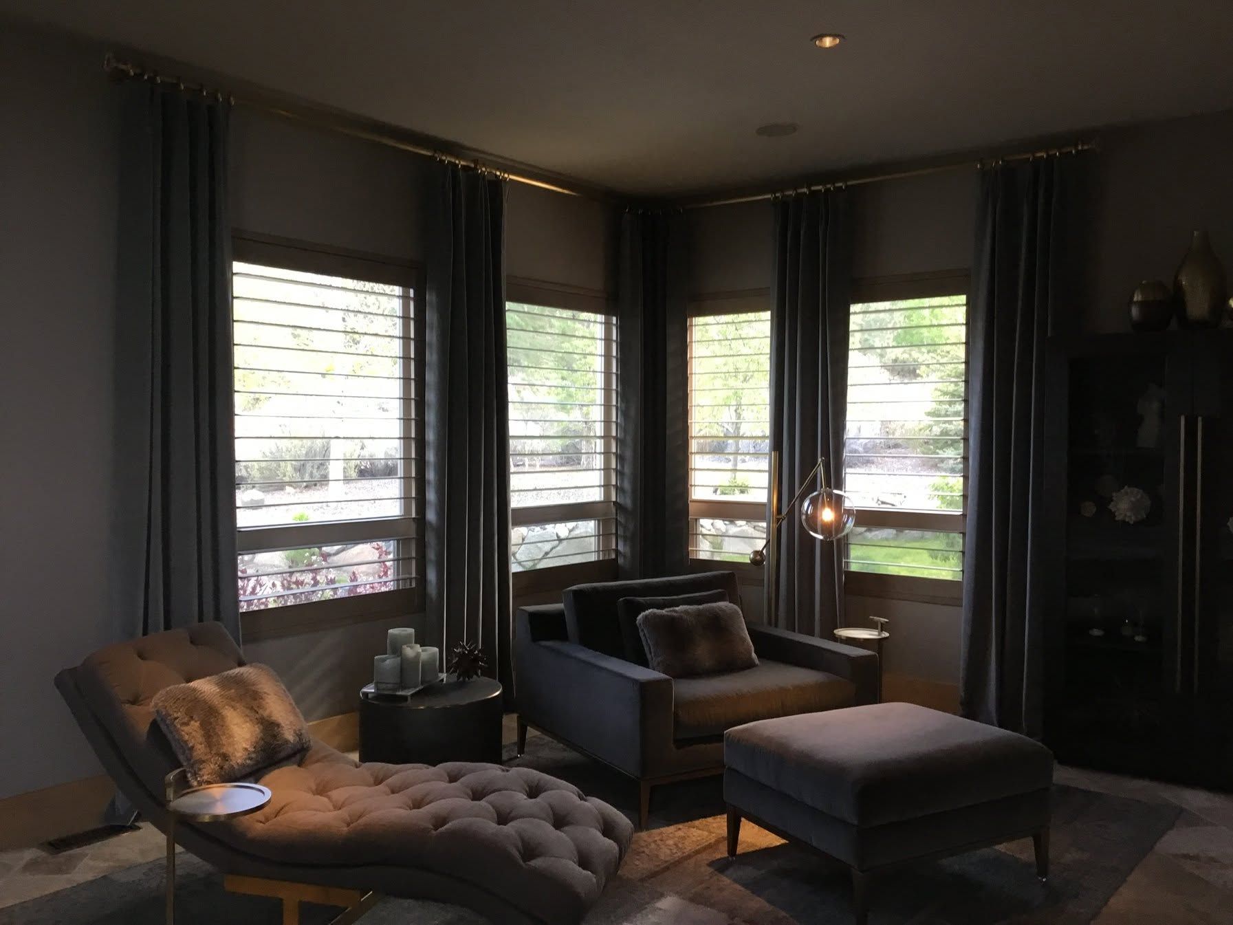 Dark modern living room with tufted chaise, armchair, ottoman, and tall windows overlooking greenery