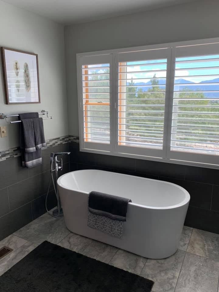 Modern bathroom with freestanding tub, black fixtures, gray tile, and window shutters