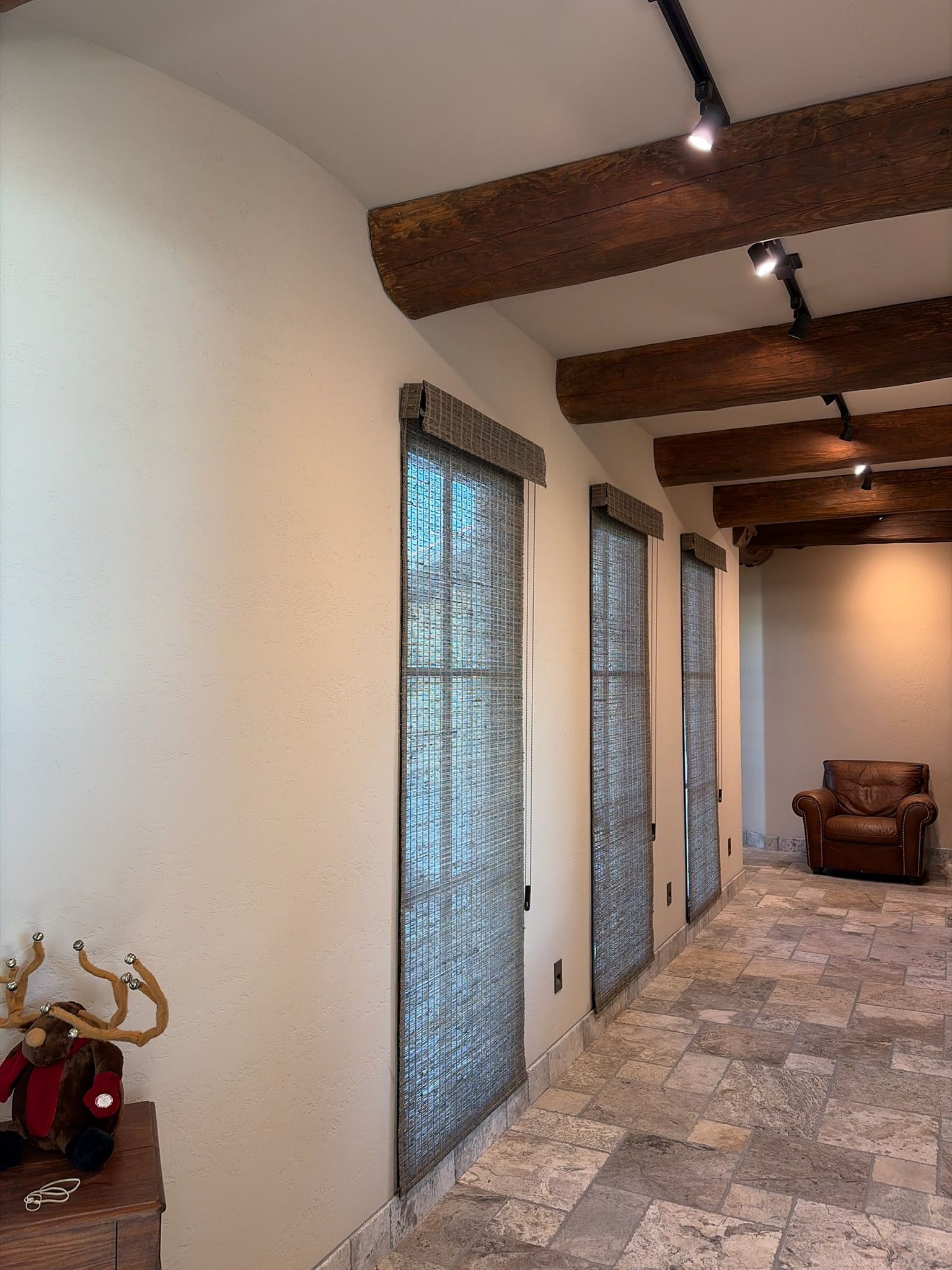 Hallway with stone floor, exposed wood beams, tall narrow windows, and a brown chair at the far end