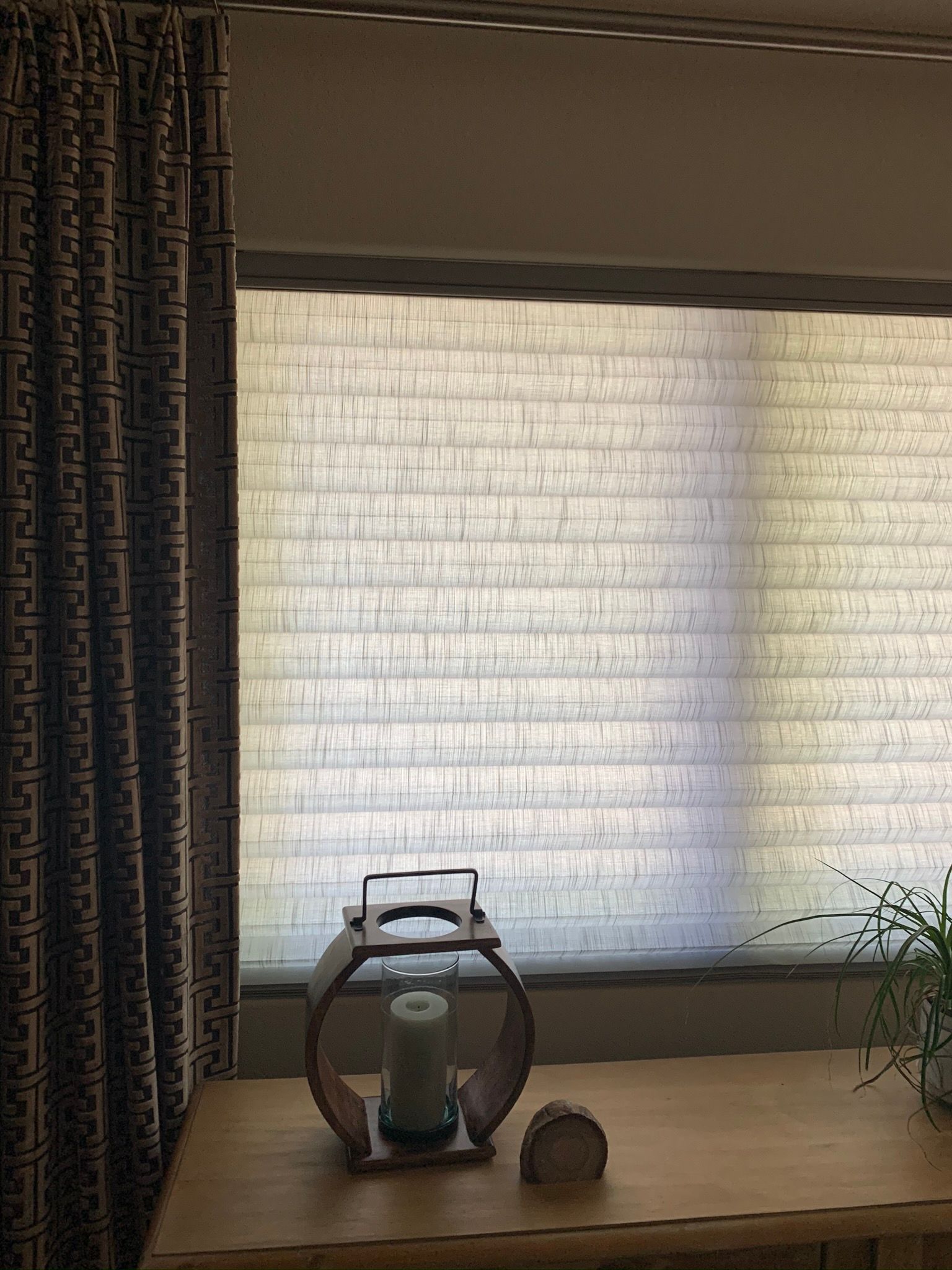 A small lantern on a wooden table beside a sunlit window with white blinds and dark curtains.