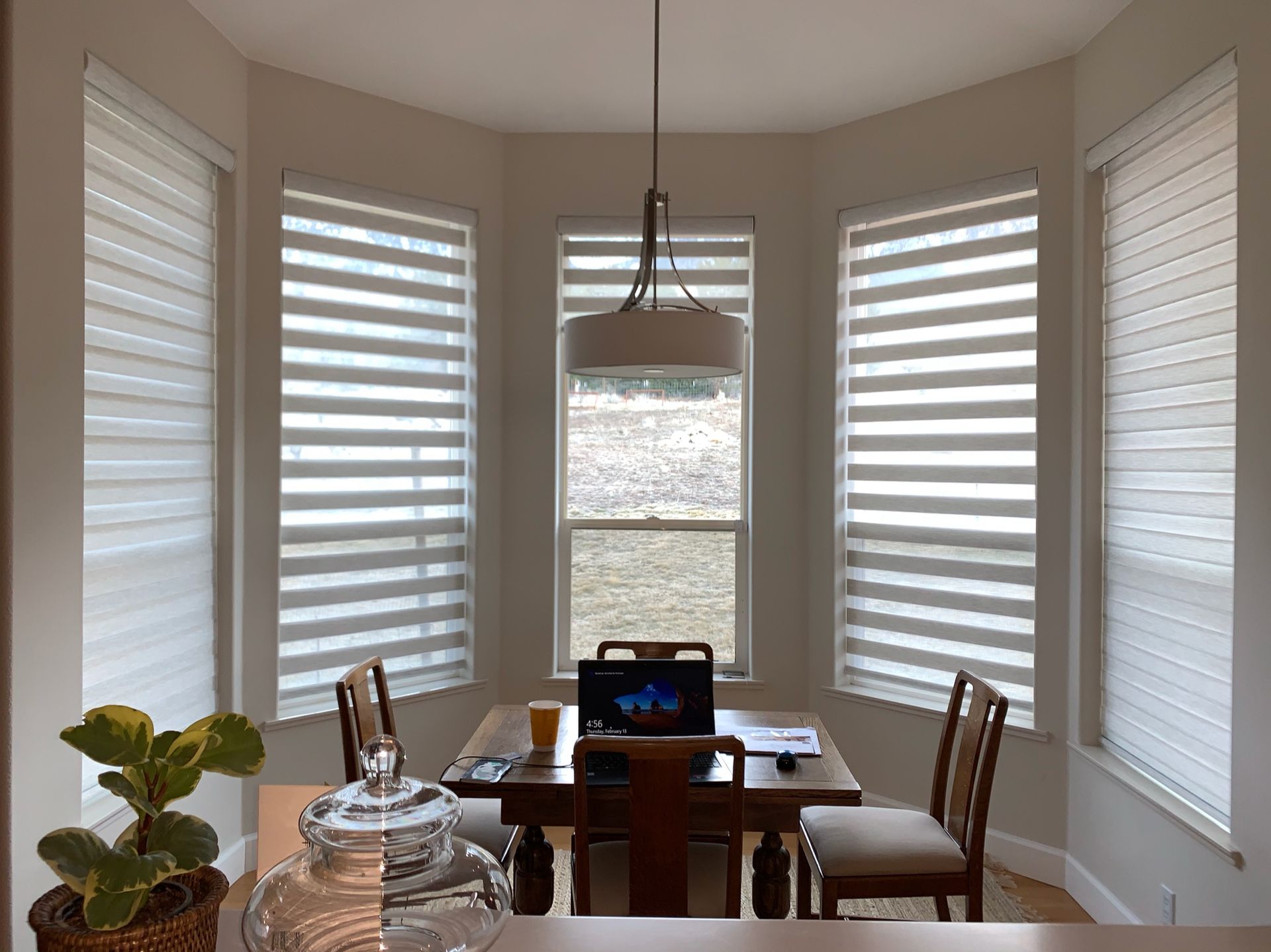 Bright dining nook with a table, chairs, laptop, and large white blinds around a corner window