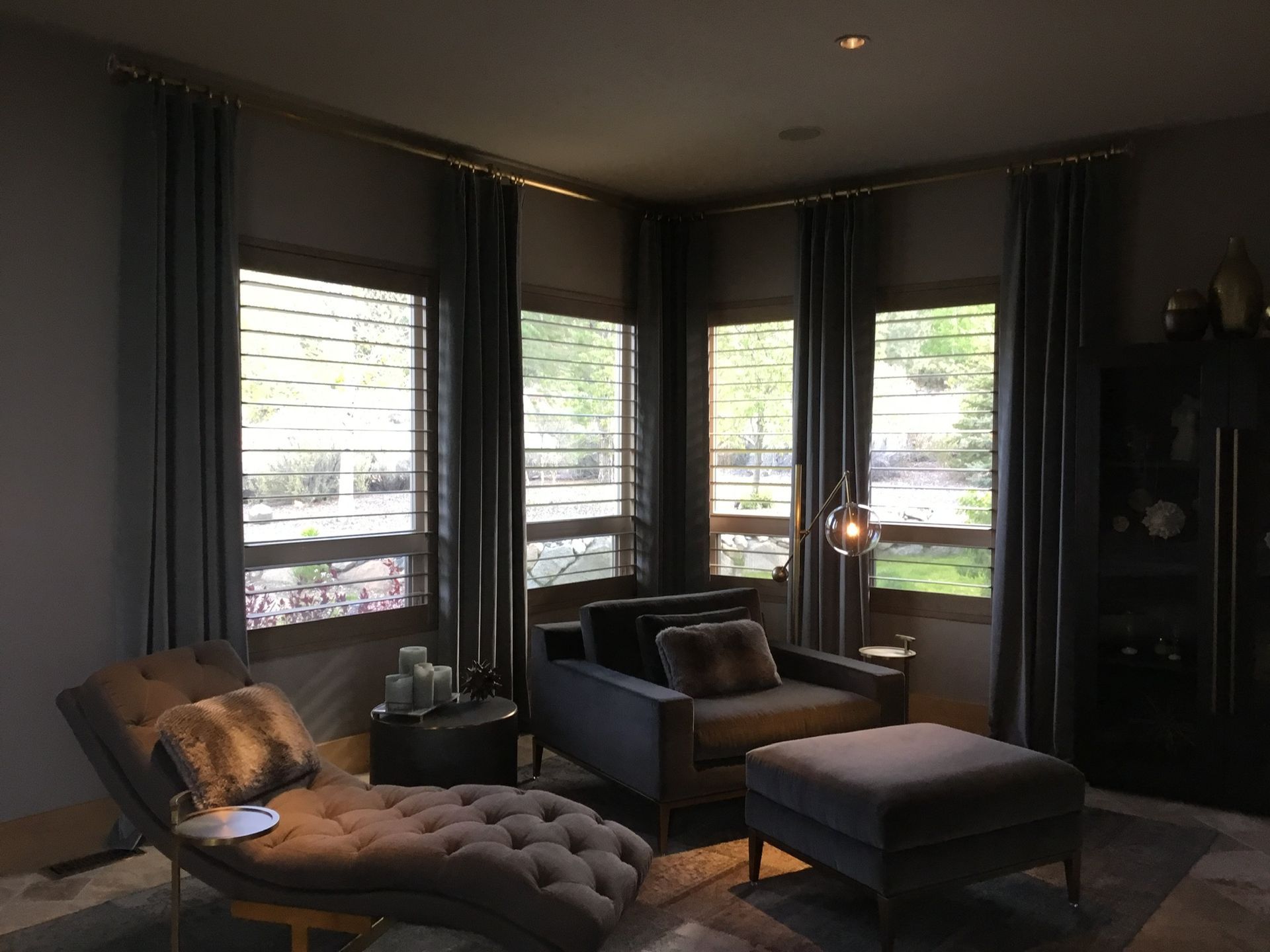 Modern dark living room with large windows, tufted chaise, armchair, ottoman, and muted gray decor.