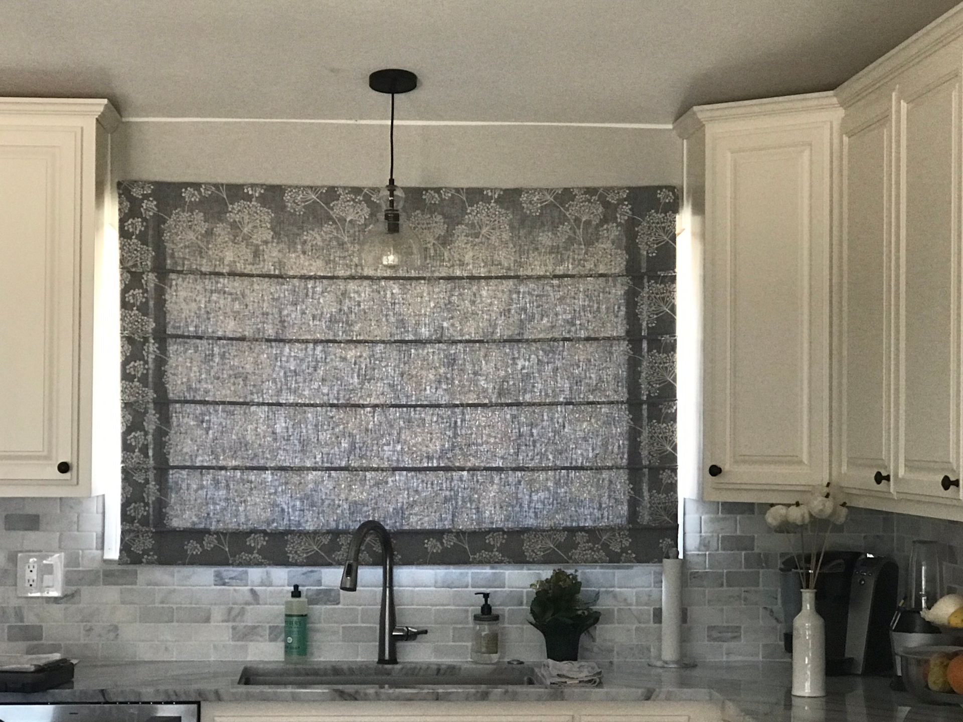 Kitchen sink with white cabinets, tiled backsplash, and a window covered by a patterned roman shade.