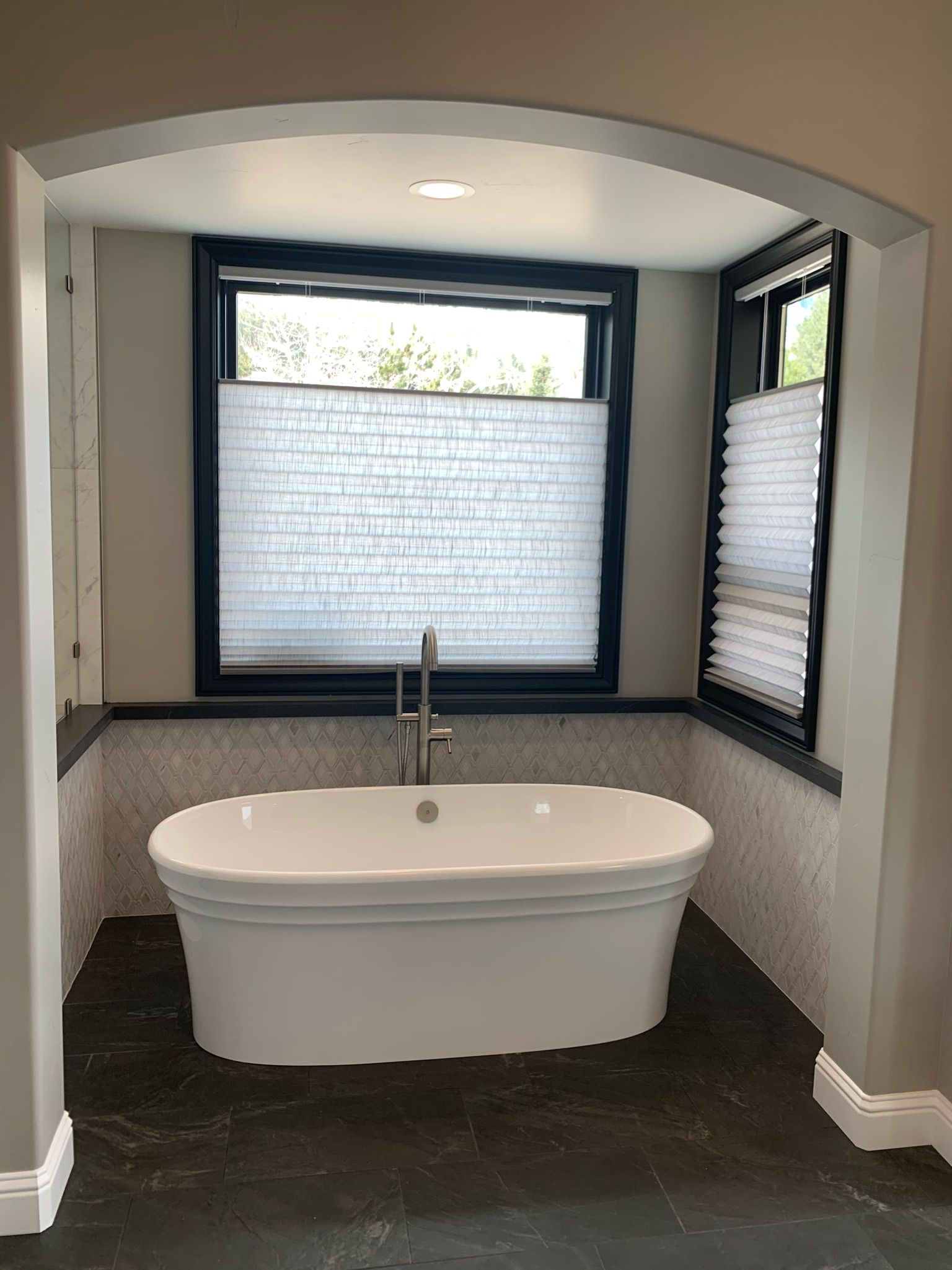 Modern bathroom with a freestanding tub beneath a window and arched alcove, dark floor and blinds.