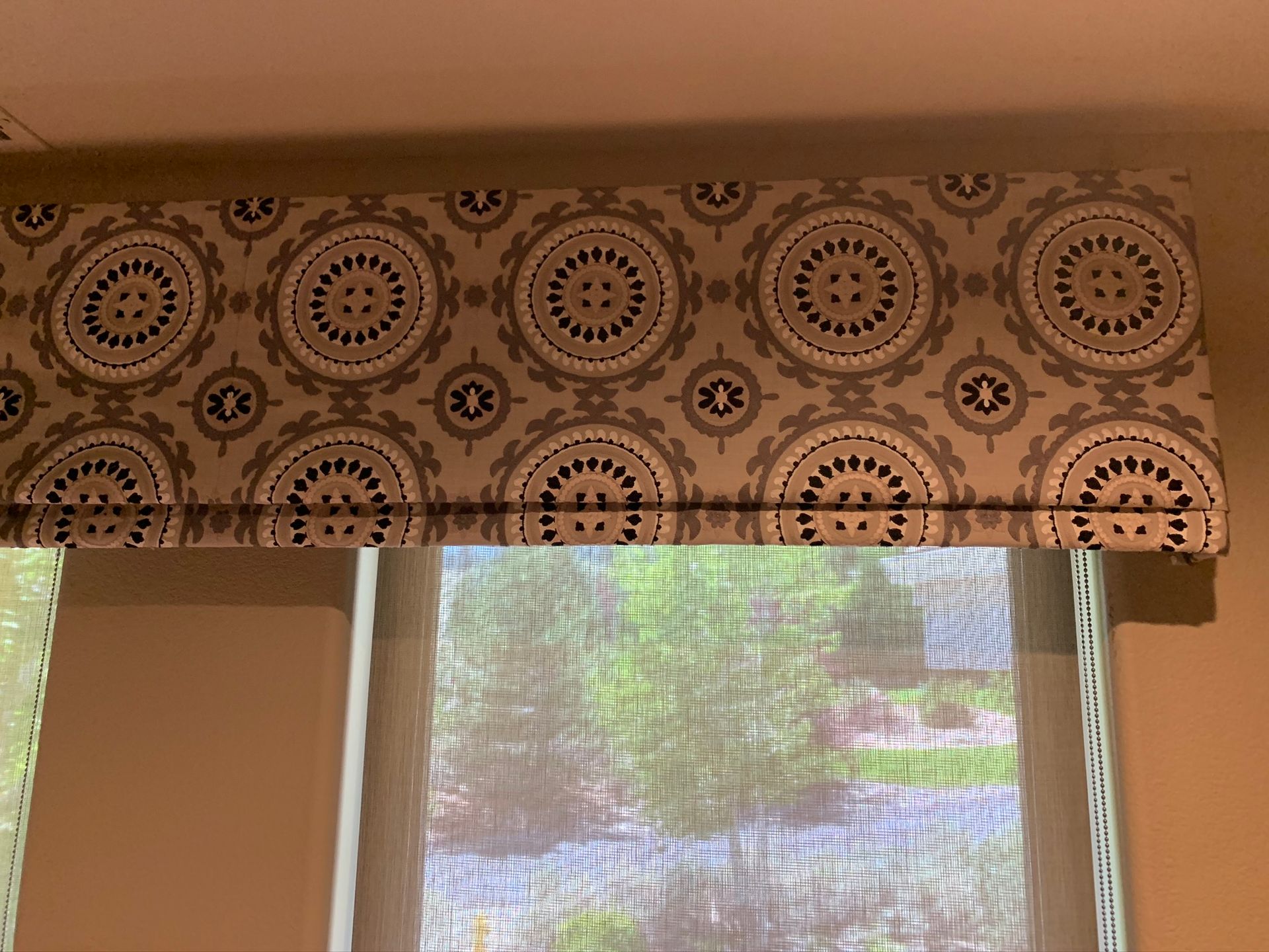 Patterned window valance above a window with a sheer curtain and outdoor greenery beyond