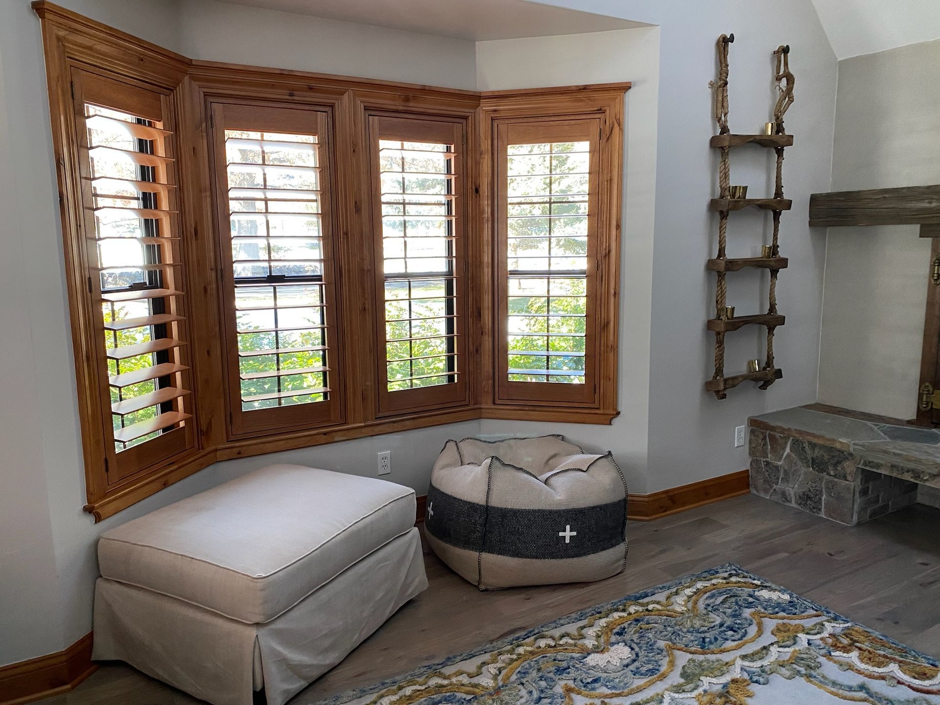 Cozy corner with bay windows, white ottoman, gray pet bed, wall ladder shelf, and stone fireplace on a patterned rug