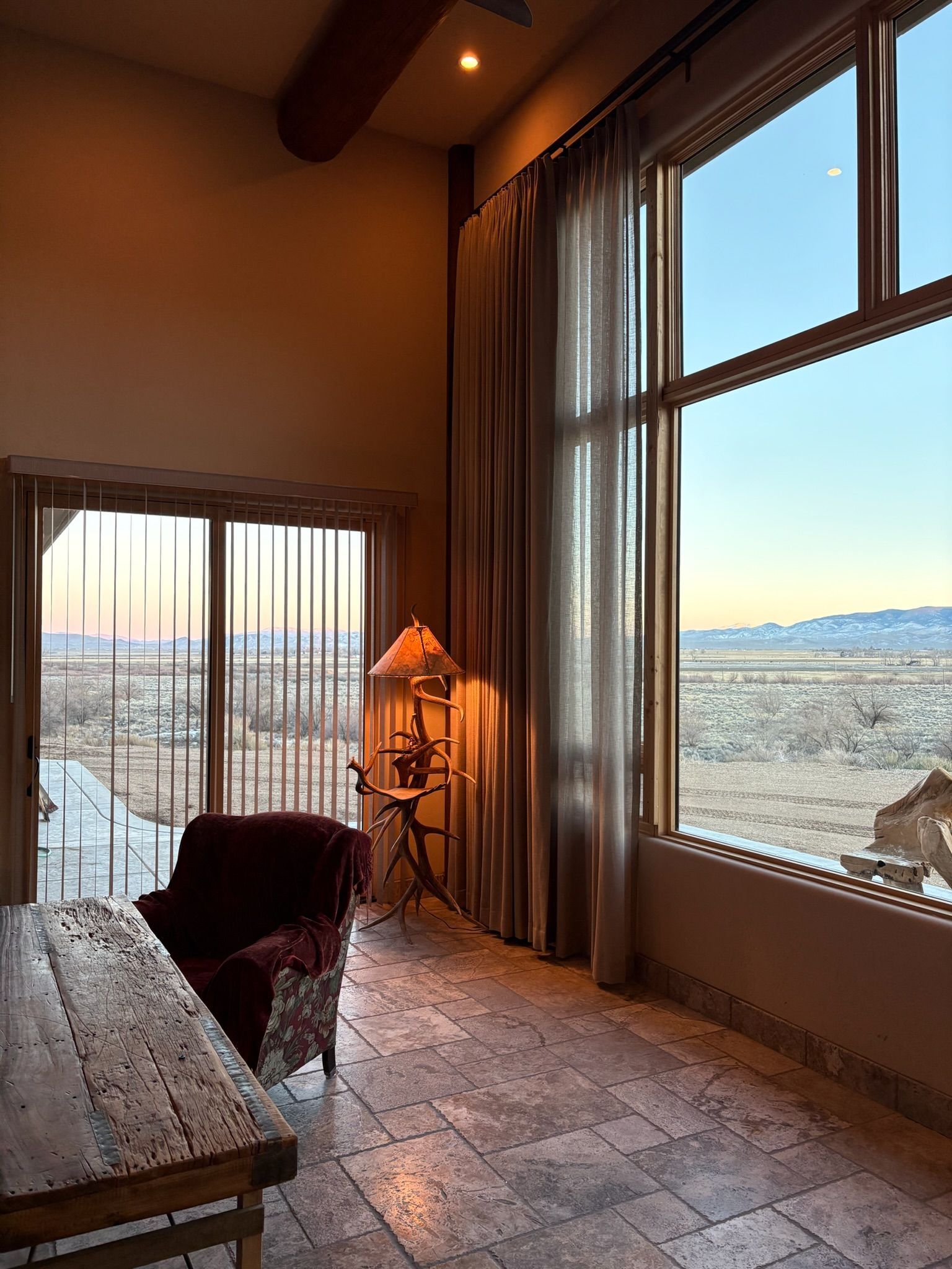 Sunlit room with large windows overlooking snowy mountains and a distant cityscape; chair by the window.