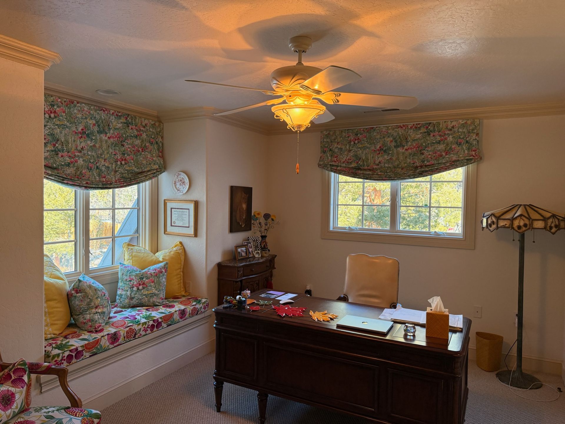 Bright home office with a dark wood desk, floral window seats, and a lit ceiling fan under patterned valances.