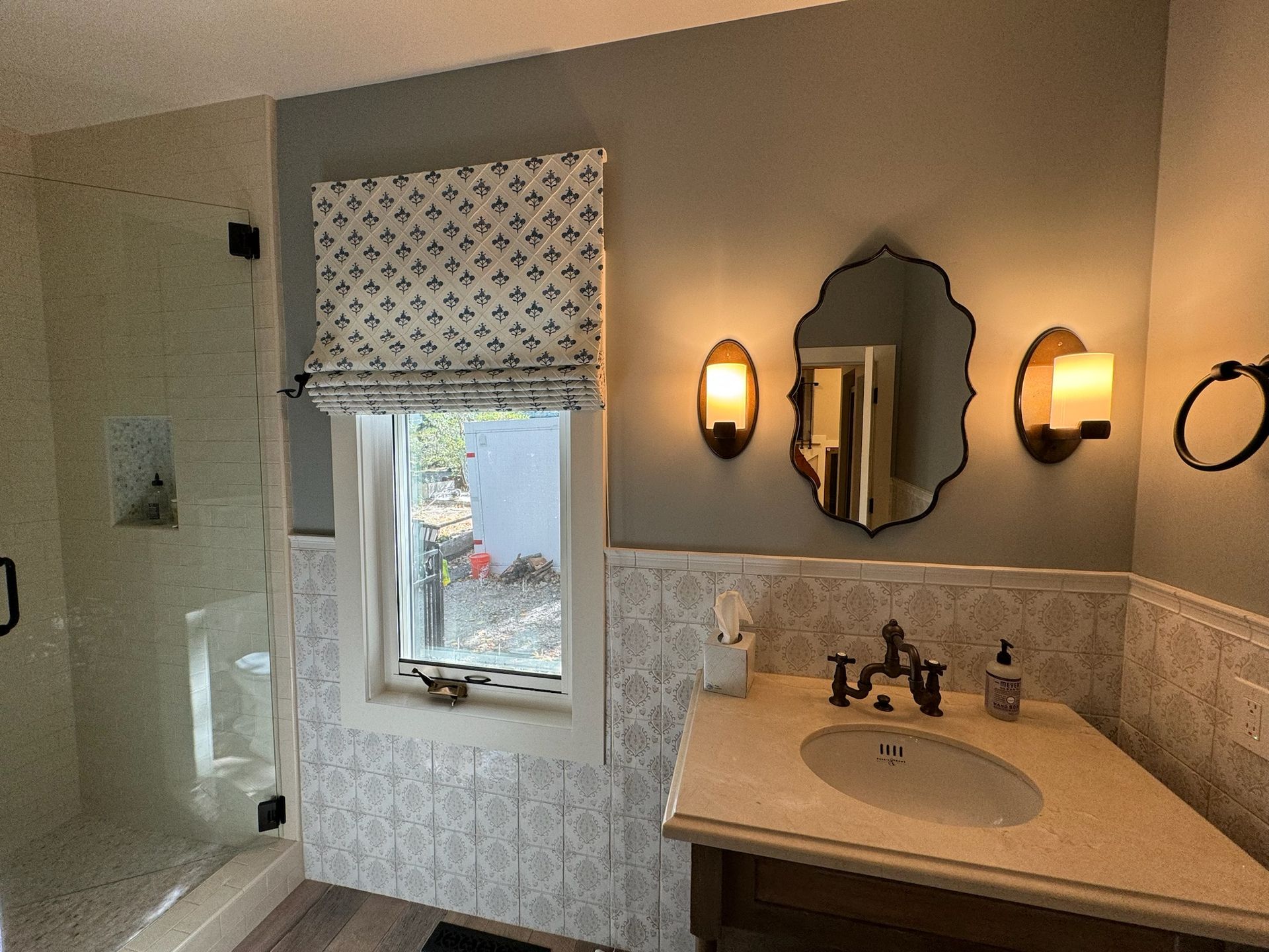 Bright bathroom with window, glass shower, vanity sink, mirror, and wall sconces