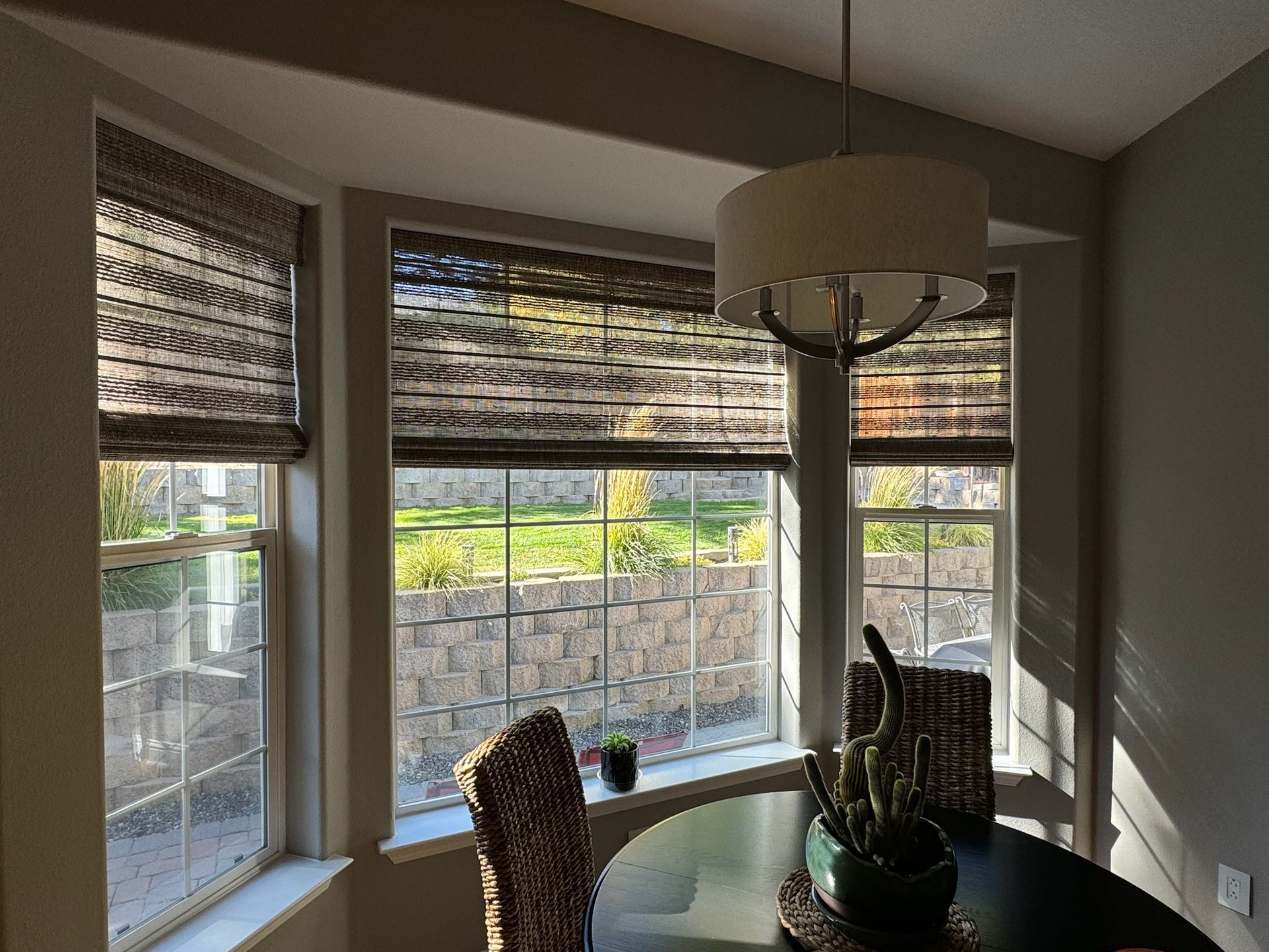 Bay window dining nook with woven shades, chair, and pendant light in a sunlit room
